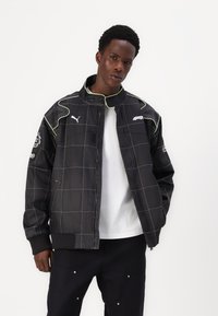 Black bomber jacket with grid pattern, white stitching, green accents, and PUMA logo. Features a stand-up collar and front pockets.