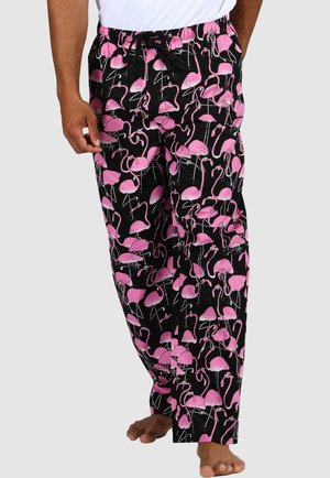 Mann trägt schwarze Pyjamahosen mit pinkem Flamingo-Muster und weißem Hemd, barfuß vor einfarbigem Hintergrund.