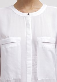 Vit, lättviktig skjorta med knappstängning och två bröstfickor med horisontella öppningar. Slät textur och klassisk kragsdesign.