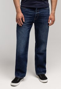 Mężczyzna w ciemnoniebieskich prostych dżinsach i czarnych sneakersach z białymi podeszwami na jednolitym jasnym tle.