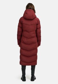 Cappotto invernale maroon con cappuccio, caratterizzato da una silhouette lunga, un motivo a chevron e polsini elasticizzati. Indossato sopra pantaloni neri e stivali.