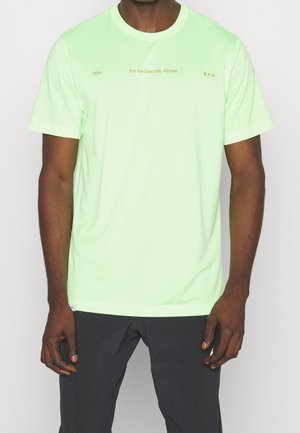 Mężczyzna w jasnozielonej koszulce Nike z napisem „Nike For the Everyday Athlete S.7-2” i czarnych spodniach na jednolitym tle.