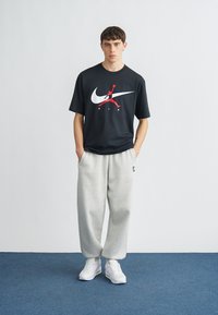 Μαύρο κοντομάνικο μπλουζάκι με μεγάλο λευκό λογότυπο Nike και κόκκινο γραφικό Jumpman, συνδυασμένο με γκρι φόρμα και λευκά αθλητικά παπούτσια.