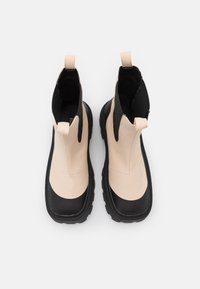 Botas de tornozelo em bege e preto com acabamento brilhante, painéis laterais elásticos e solas de borracha texturizadas. Dois puxadores para facilitar o calçar.