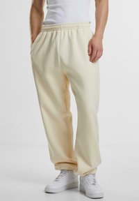 Hombre vestido con pantalones de chándal elásticos color blanco roto y zapatillas blancas, de pie con una mano en el bolsillo frente a un fondo liso.