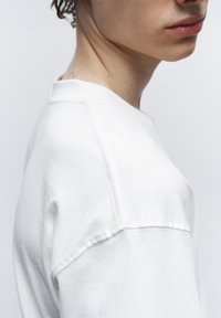 Camiseta blanca de algodón con escote redondo, costuras visibles y un corte relajado. La tela tiene una textura suave.