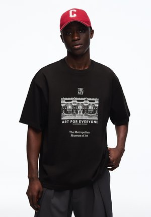 Tânăr purtând o șapcă roșie și un tricou negru cu o grafică alb-negru a clădirii Metropolitan Museum of Art și textul „Artă pentru toți.”