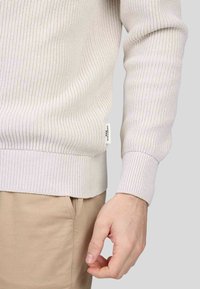 Pull en maille côtelée beige clair avec un tissu texturé, des poignets et un ourlet ajustés. Étiquette visible sur la couture latérale. Assorti avec un pantalon beige clair.