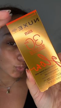 Personne tenant une boîte dorée du fluide ensoleillé NUXE Sun SPF 30 avec des ongles brillants et un collier.