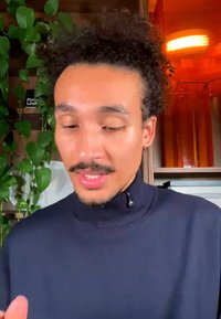 Homme aux cheveux bouclés et à la moustache portant un col roulé foncé, parlant à l’intérieur avec des plantes et des étagères en arrière-plan.