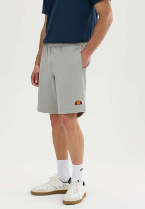 Mann trägt hellgraue Sportshorts mit kleinem Logo, weiße Socken, weiße Sneakers und ein dunkelblaues Hemd, steht mit einer Hand in der Tasche.