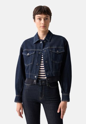 Person trägt eine dunkelblaue kurze Jeansjacke über einem gestreiften Hemd und passenden dunkelblauen Jeans, steht vor einem einfarbigen Hintergrund.
