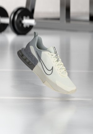 Svetlošedá a krémová teniska Nike Air Max s vrchnou časťou z sieťoviny vznášajúca sa nad lesklou podlahou telocvične, rozmazané závažiarské vybavenie v pozadí.