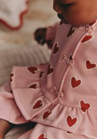 Rosa bomullsoutfit med röda hjärtmönster och vita detaljer. Har små rosetter på knapparna och en volangkant.