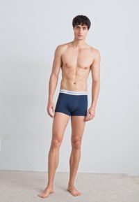 Marineblaue Baumwollboxershorts mit einem weißen elastischen Bund, auf dem der Markenname "GANT" aufgedruckt ist. Das Modell steht auf einem hellen Teppich.