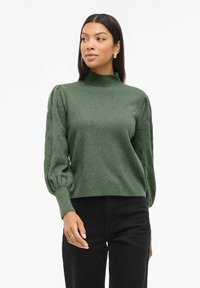 Maglione verde con colletto alto, polsini a coste e maniche lunghe a sbuffo. Materiale testurizzato, design leggermente corto. Indossato con pantaloni neri.