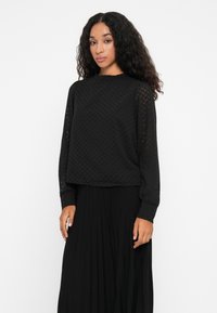 Black long-sleeve top with a textured zigzag pattern, round neckline, and relaxed fit, paired with a flowing black pleated skirt.