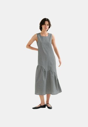 Kvinde iført en sort-hvid ternet ærmeløs midikjole med asymmetrisk flæsekant, kombineret med sorte Mary Jane-flade sko.