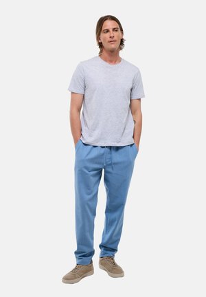 Homme debout, les mains dans les poches, portant un t-shirt gris clair, un pantalon bleu et des baskets beiges sur un fond blanc uni.