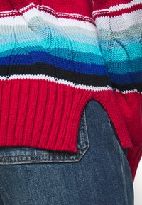 Pull en maille rayé rouge et bleu avec ourlet côtelé. Présente un dégradé de rouge à bleu, mettant en valeur des points texturés en tricot.