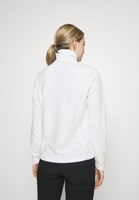 Weißes Fleece-Jackett mit hohem Kragen, langen Ärmeln und seitlichen Nähten. Weiche Textur mit einem einfachen, klaren Design, getragen über einer schwarzen Hose.