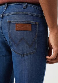 Granatowe dżinsy o slim fit, z charakterystyczną brązową skórzaną metką Wrangler na tylnej kieszeni oraz kontrastującym żółtym szwem.