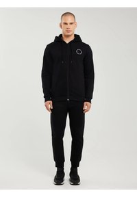 Bărbat purtând hanorac negru cu fermoar și text circular alb pe piept, pantaloni negri sport și adidași negri, stând în picioare pe un fundal simplu și deschis la culoare.