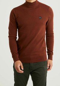 Brązowy sweter z golfem wykonany z miękkiego dzianinowego materiału, z żebrowanym kołnierzem i mankietami oraz niewielką czarną naszywką z logo na piersi.