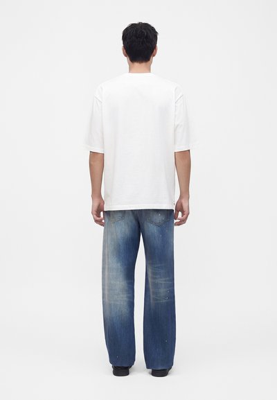 Άνδρας με πλάτη, φοράει απλό λευκό oversized κοντομάνικο μπλουζάκι και φαρδιά ξεβαμμένα μπλε τζιν με μαύρα παπούτσια σε λευκό φόντο.