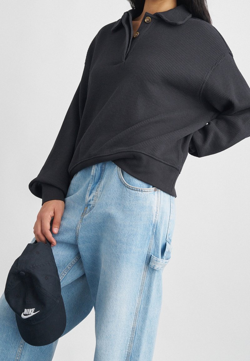 Personne portant un pull en maille gaufrée noir avec des boutons, un jean bleu clair, tenant une casquette Nike noire à côté d'elle.