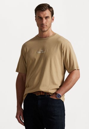 Beige pamut póló kerek nyakkal, rövid ujjakkal és hímzett logóval a mellrészen. Sötét farmernadrággal és fonott övvel viselve.