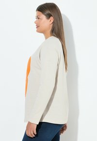 Beige långärmad t-shirt med en orange hjärt-design framtill. Tillverkad av mjukt tyg, har den en avslappnad passform och rund halsringning.