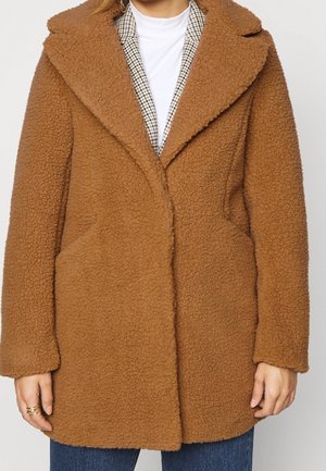 Brun teddy-frakke med bred krave båret over ternet blazer og hvid top, kombineret med blå jeans, viser den forreste del af overkroppen og hænderne.