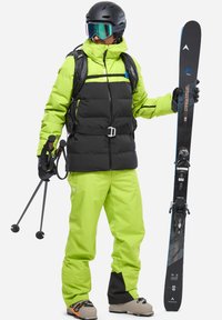 Ski-Ausrüstung mit einer leuchtend grünen und schwarzen, isolierten Jacke und Hose, Skistöcken und einem schwarzen Skihelm mit reflektierenden Goggles.