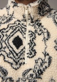 Fleece pullover med en kvart-lynlås, der har en cremefarvet base og sorte geometriske mønstre, tekstureret stof og et højt kravedesign.