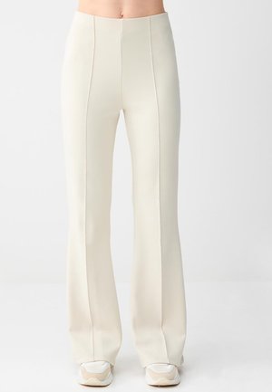 Persoon die een crème-kleurige broek met hoge taille en wijde pijpen draagt, met naaddetails, gecombineerd met witte en beige sneakers, staand voor een effen achtergrond.