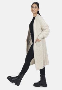 Larga chaqueta de punto abierta color beige con hombros caídos y bolsillos, llevada sobre una camiseta negra y leggings negros, combinada con botas de tobillo negras con cordones.