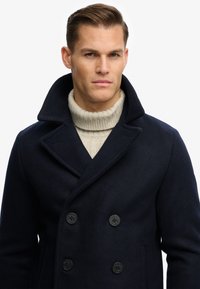 Man draagt een marineblauwe dubbelgeknoopte jas met een brede kraag over een crèmekleurige kabelgebreide coltrui, kijkt vooruit met een neutrale uitdrukking.