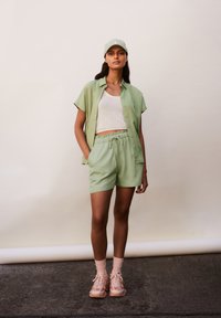 Lichtgroen shirt met korte mouwen, witte geribbelde tanktop, bijpassende shorts met koord en roze sneakers met lichtroze sokken. Een pet maakt de look compleet.