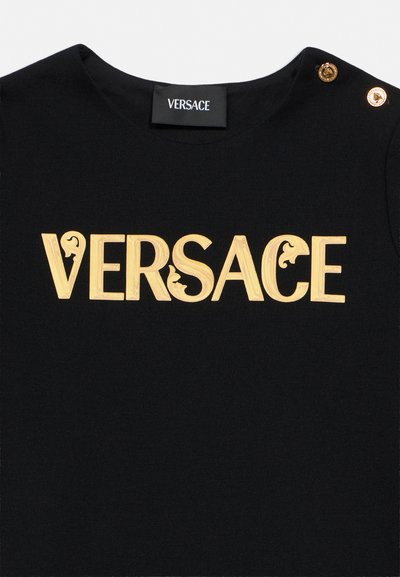 Haut noir Versace avec un logo doré en relief sur la poitrine et deux boutons dorés sur l'épaule.
