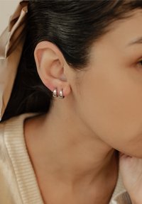 Srebrne kolczyki w kształcie kół, umieszczone na uchu modelki, z gładką, odbijającą powierzchnią i zwężającym się kształtem.