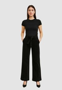 Top nero a coste con maniche corte abbinato a pantaloni in corda neri a gamba larga con vita con cintura. La modella sta in piedi con le mani nelle tasche.