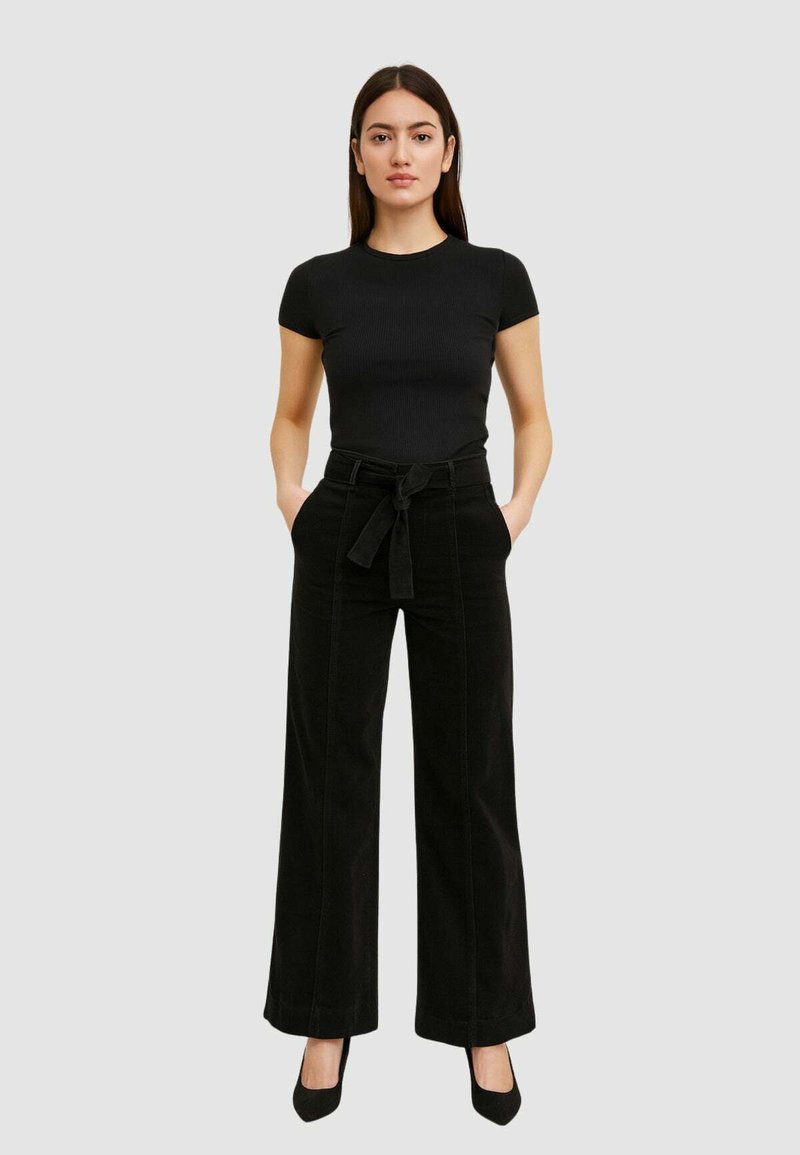 Top nero a coste con maniche corte abbinato a pantaloni in corda neri a gamba larga con vita con cintura. La modella sta in piedi con le mani nelle tasche.