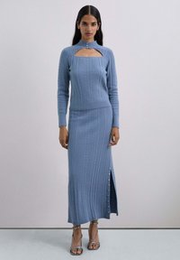 Mujer con un suéter de punto en cable azul claro con corte en el pecho y falda larga a juego con una abertura lateral y detalles de botones de perlas, tacones plateados.