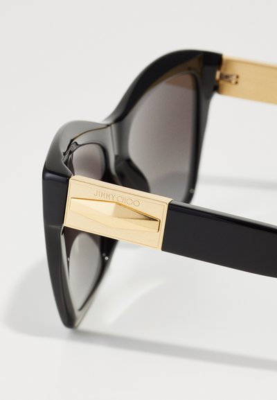Lunettes de soleil noires avec verres gris, dotées d'une monture en plastique audacieuse et de détails en métal doré sur les branches, gravées avec le nom de la marque.