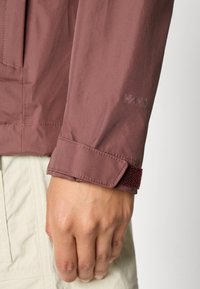 Hand rustend aan de zijde, met mauve jasje waarvan de mouwen een verstelbare manchet hebben, beige broek zichtbaar op de achtergrond.