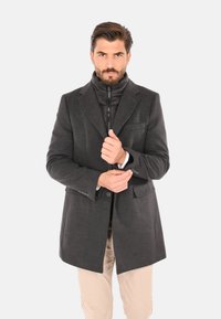 Uomo con barba che sistema un bottone su un cappotto grigio scuro sopra una giacca nera con cerniera, indossando pantaloni beige, in piedi davanti a uno sfondo bianco uniforme.