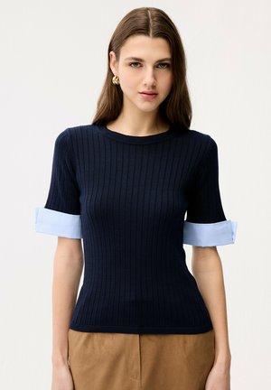 Junge Frau mit langen braunen Haaren, die goldene Creolen und ein marineblaues, geripptes Kurzarmshirt mit hellblauen umgeschlagenen Ärmelabschlüssen sowie braune Hosen trägt.