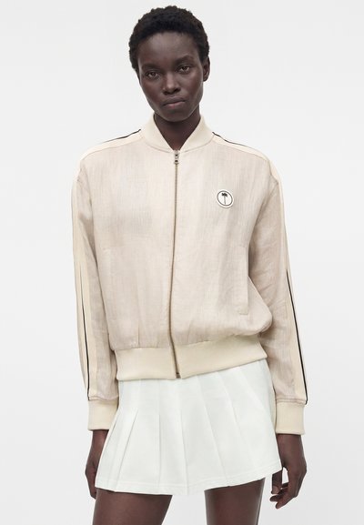 Γυναίκα που φοράει ελαφρώς μπεζ bomber μπουφάν με φερμουάρ και μαύρες ρίγες στα μανίκια και λευκή πλισέ φούστα σε λευκό φόντο.