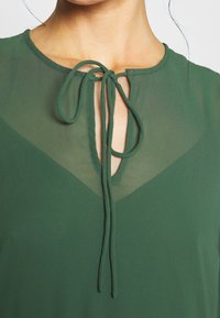 Blusa verde con cuello de lazo y un panel transparente en el frente. La tela parece suave y tiene una textura ligera.